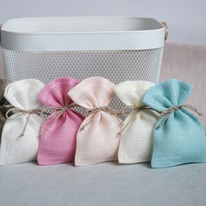 May include: Five small fabric gift bags in different colors. The bags are made of a natural linen-like material and are tied with twine. The bags are arranged in a row on a white surface.