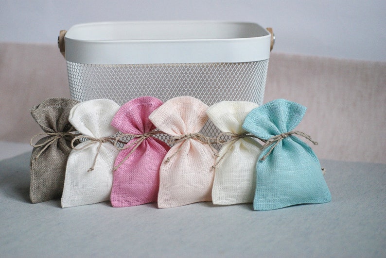 May include: Five small fabric gift bags in different colors. The bags are made of a natural linen-like material and are tied with twine. The bags are arranged in a row on a white surface.