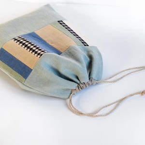 May include: A light blue drawstring bag with a patchwork design of blue, beige, and black checkered fabric. The bag has a drawstring closure and is made of a soft, natural fabric.