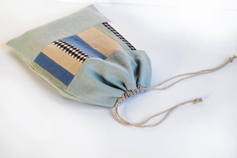 May include: A light blue drawstring bag with a patchwork design of blue, beige, and black checkered fabric. The bag has a drawstring closure and is made of a soft, natural fabric.