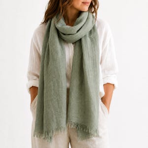 Puede incluir: Una bufanda de color verde salvia con una apariencia tejida y texturizada y bordes deshilachados. La bufanda está colocada holgadamente alrededor del cuello, con un acabado mate suave y una forma rectangular que cuelga al frente.