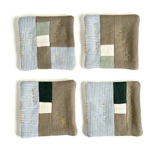 May include: Four square fabric coasters with a patchwork design. Each coaster features a combination of light blue striped fabric, olive green, white, and brown fabric. Gold embroidery accents the design.