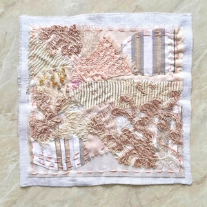 Può includere: Opera d'arte tessile con un design patchwork. L'opera presenta varie trame e colori, tra cui sfumature di rosa, beige e bianco. Il pezzo è incorniciato con un bordo bianco e presenta cuciture e abbellimenti intricati, creando un'estetica unica e artigianale.