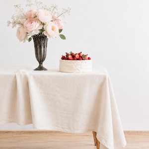 Könnte beinhalten: Ein runder, weiß glasierter Kuchen, der mit frischen roten Erdbeeren und Himbeeren belegt ist, steht neben einer dunklen Metallvase mit hellrosa Rosen und weißem Schleierkraut auf einer beigen Leinentischdecke, die einen Holztisch bedeckt.
