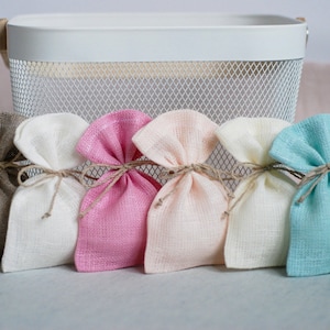 May include: Six small fabric gift bags in various colors, including beige, white, pink, peach, cream, and light blue. The bags are tied with twine and are arranged in a row on a gray surface.