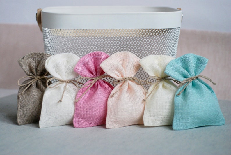 May include: Six small fabric gift bags in various colors, including beige, white, pink, peach, cream, and light blue. The bags are tied with twine and are arranged in a row on a gray surface.