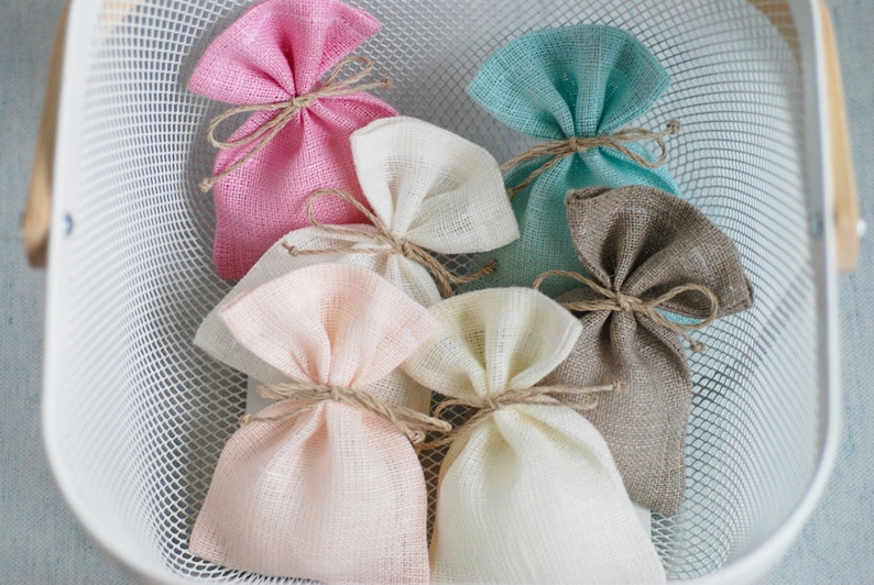 May include: A white wire basket filled with six small burlap gift bags. The bags are tied with twine and come in a variety of colors, including pink, beige, light blue, and brown.