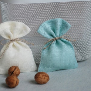 May include: Two small fabric gift bags, one white and one light blue, tied with twine. The bags are sitting on a light blue surface with two walnuts in front of them.