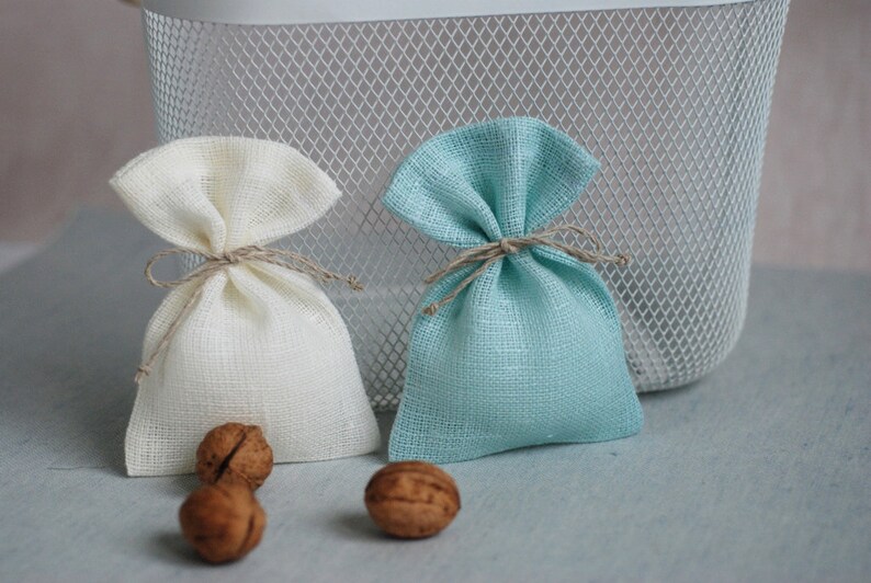 May include: Two small fabric gift bags, one white and one light blue, tied with twine. The bags are sitting on a light blue surface with two walnuts in front of them.