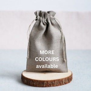 Può includere: Un sacchetto grigio con coulisse con il testo "MORE COLOURS available" è esposto su una piccola fetta di legno. La borsa è realizzata in un tessuto strutturato e ha una parte superiore arricciata con chiusura a coulisse.