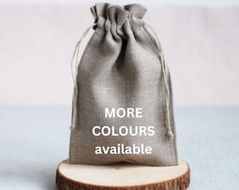 May include: A gray drawstring bag with the text "MORE COLOURS available" is displayed on a small wooden slice. The bag is made of a textured fabric and has a gathered top with a drawstring closure.