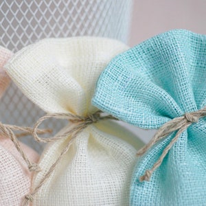 May include: Three small fabric gift bags in pink, white, and light blue. The bags are made of a woven material and are tied with twine.