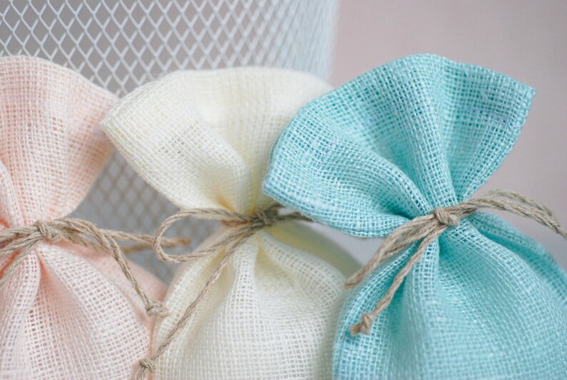 May include: Three small fabric gift bags in pink, white, and light blue. The bags are made of a woven material and are tied with twine.