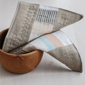 Peut inclure: Deux carrés de tissu pliés avec un motif patchwork dans des tons beige, gris et blanc. Les carrés de tissu sont en lin et ont un aspect cousu à la main. Les carrés sont posés sur un bol en bois.