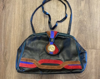 May include: A black leather handbag with a brown, red, and blue suede trim. The bag has a gold-toned circular accent on the front.