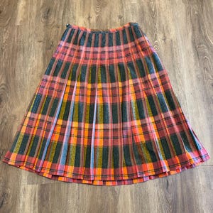 May include: A pleated midi skirt featuring a plaid pattern in shades of dark green, bright orange, and muted pink. The skirt has a fitted waistband and a flared silhouette with vertical pleats running from the waist to the hemline.