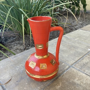May include: A red ceramic pitcher with a gold geometric design. The pitcher has a round body and a tall, narrow spout. It has a handle on the side.