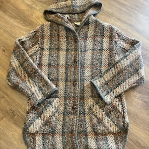 May include: A hooded cardigan featuring a textured plaid pattern in shades of brown, gray, and cream. The garment has long sleeves, a button-front closure with four dark brown buttons, and two front patch pockets with a visible knit construction.