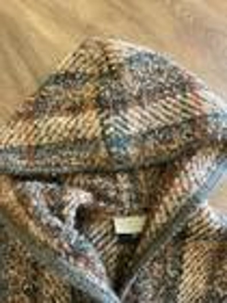 May include: A hooded jacket featuring a thick, textured knit fabric with a multi-colored plaid pattern in shades of brown, tan, grey, and rust. The garment includes a hood, a front opening, and a small rectangular label sewn inside the neckline.