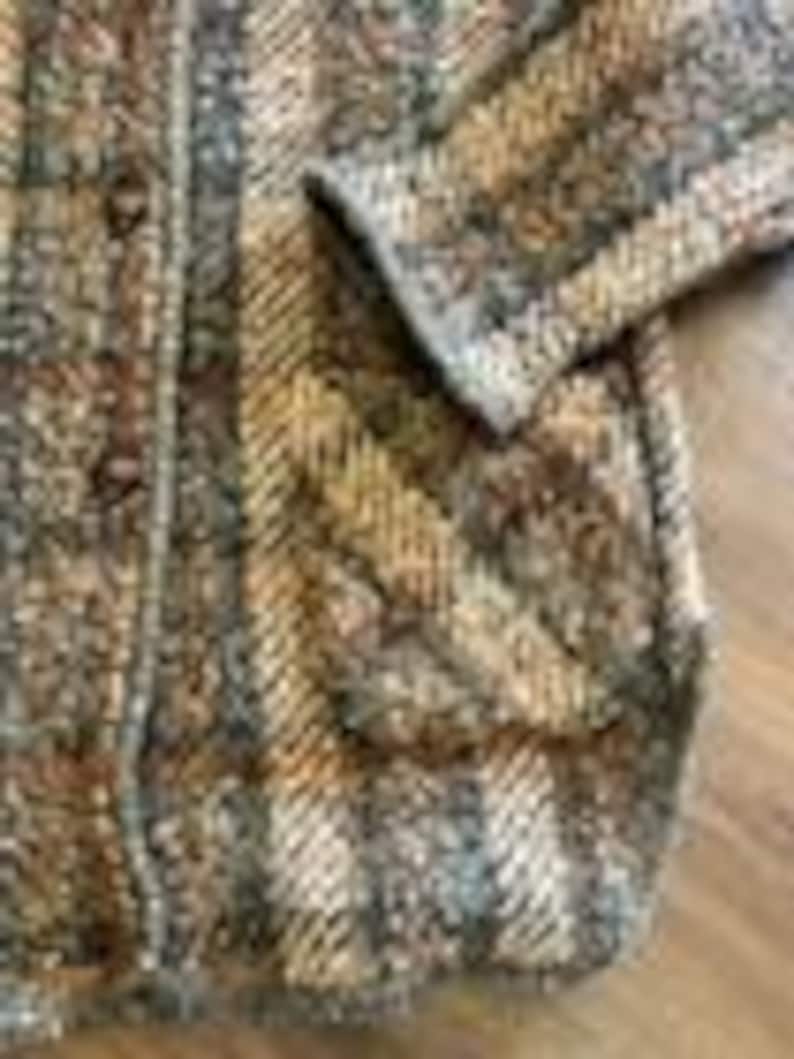 May include: A long-sleeved cardigan featuring a textured knit pattern in shades of brown, tan, gray, and cream. The garment includes a vertical striped design, a button-down front closure with visible buttons, and a patch pocket on the front side.