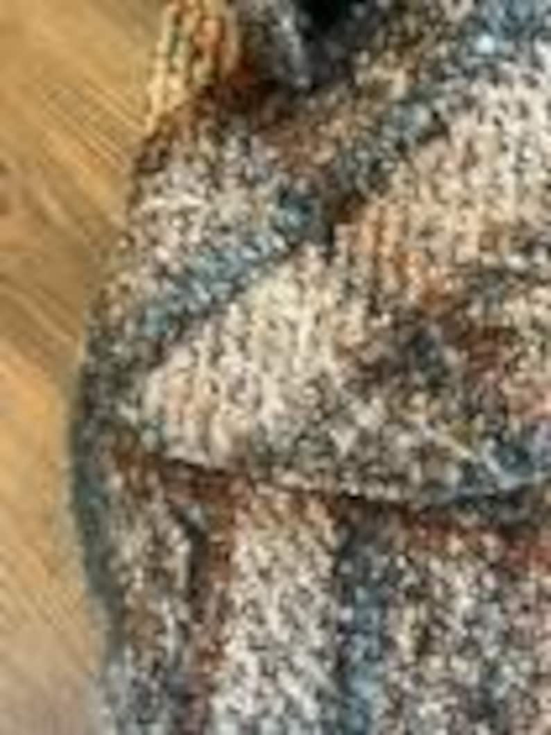 May include: A chunky knit textile featuring a variegated color palette of brown, tan, cream, and slate blue yarns. The fabric displays a thick, textured weave with visible loops and twisted fibers, creating a dense and multi-tonal appearance throughout.