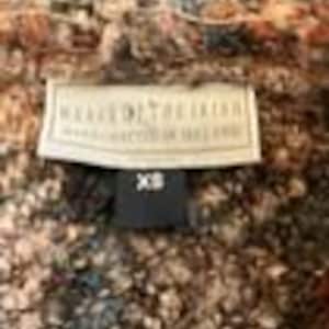 May include: A textured, multi-colored knit garment featuring a cream-colored rectangular label with the text "WEAVE OF THE IRISH" and "MANUFACTURED IN IRELAND" in black, with a smaller black tag below it displaying the size "XS" in white text.