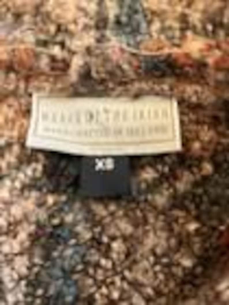 May include: A textured, multi-colored knit garment featuring a cream-colored rectangular label with the text "WEAVE OF THE IRISH" and "MANUFACTURED IN IRELAND" in black, with a smaller black tag below it displaying the size "XS" in white text.