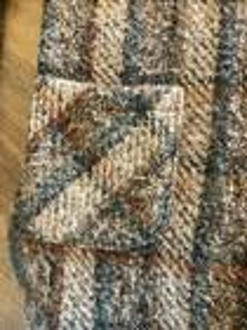 May include: A textured knit fabric featuring a vertical striped pattern in shades of brown, tan, gray, and white. The material displays a thick, woven construction with a visible pocket integrated into the striped design, creating a multi-dimensional surface.