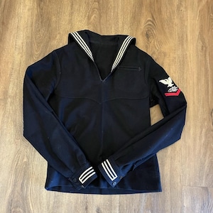 May include: A dark navy blue long-sleeve sailor jumper featuring a wide collar with three white stripes, matching white stripes on the cuffs, and a white eagle emblem with a red chevron patch on the left sleeve, constructed with a V-neckline.