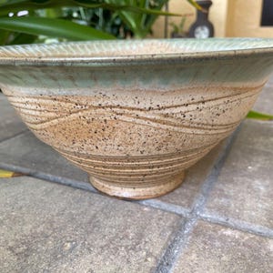 May include: A large, hand-thrown ceramic bowl with a light green glaze and brown and white speckled accents. The bowl has a wavy, textured design and a wide, rounded base.