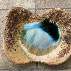 May include: A ceramic dish with a blue and green glaze. The dish is decorated with raised faces around the rim.
