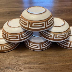 May include: Six white ceramic bowls with a brown geometric design stacked on top of each other.