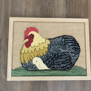 May include: A framed needlepoint artwork depicting a hen with a chick nestled under her wing. The hen is brown, yellow, and red, while the chick is white. The background is a burlap fabric.