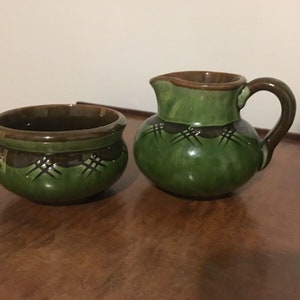 May include: A ceramic sugar bowl and creamer set featuring a glossy green glaze with dark brown rims and handles. Both pieces are decorated with a dark brown band around the upper section, accented by a repeating incised cross-hatch pattern.
