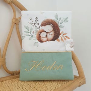 Peut inclure: Une couverture pour bébé avec une illustration à l'aquarelle de deux hérissons enlacés, sur fond de feuilles et de branches. La partie inférieure est vert clair avec le nom "Heden" brodé en fil d'or.