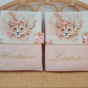 May include: Two pink fabric book covers with a lion cub illustration and floral accents. The covers have gold embroidery with the names "Hortense" and "Luna".