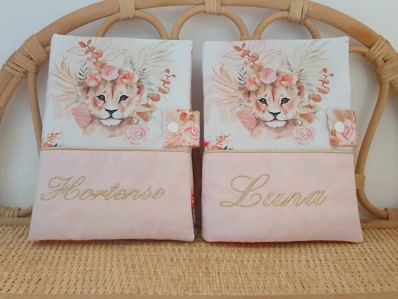 May include: Two pink fabric book covers with a lion cub illustration and floral accents. The covers have gold embroidery with the names "Hortense" and "Luna".
