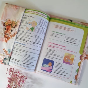May include: A pink and white fabric book cover with a giraffe illustration and floral accents. The cover has a flap with a button closure. The book's pages feature text in French about caring for babies.