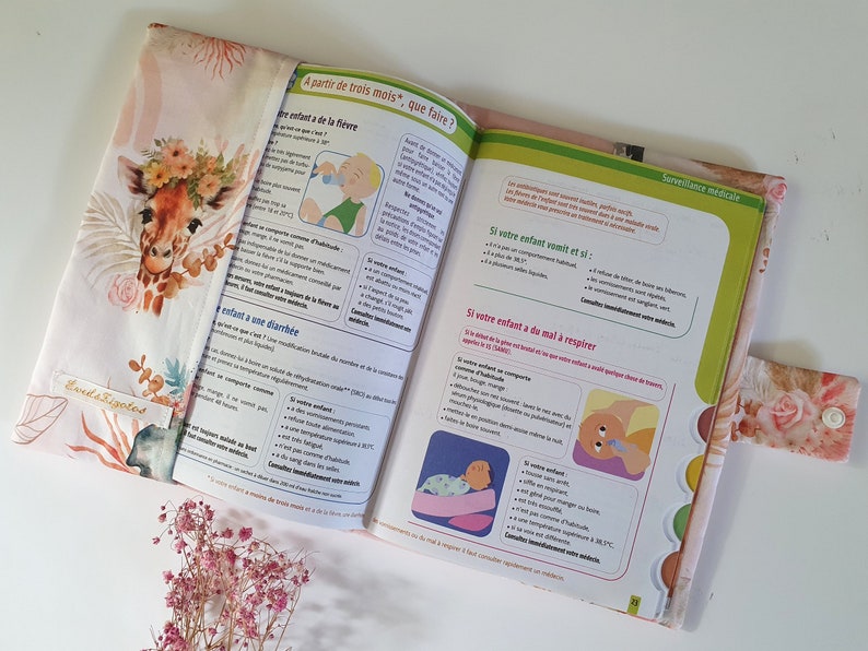 May include: A pink and white fabric book cover with a giraffe illustration and floral accents. The cover has a flap with a button closure. The book's pages feature text in French about caring for babies.