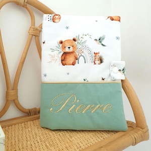 Peut inclure: Une couverture pour bébé avec un motif d'animaux de la forêt. La partie supérieure est blanche avec des illustrations d'ours, d'écureuil, d'arc-en-ciel et d'oiseaux. Le bas est vert clair avec le nom "Pierre" brodé en or.