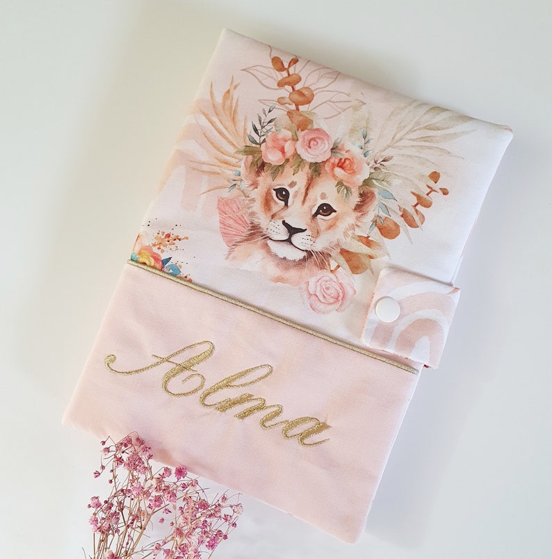 May include: A pink and white fabric book cover with a watercolor illustration of a lion cub wearing a flower crown. The cover has a gold embroidered name "Alma" on the front.