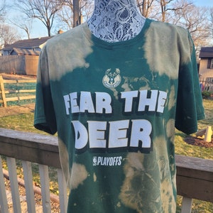 May include: Green and tan tie-dye t-shirt with the words "FEAR THE DEER" in white block letters. The shirt also features the Milwaukee Bucks logo and the word "PLAYOFFS". The t-shirt is displayed on a mannequin.
