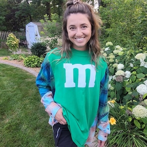May include: A green t-shirt with a large white "M" on the front. The shirt has long sleeves with a blue and green plaid pattern. The person is wearing black shorts and is standing outside in front of a garden.