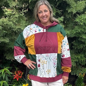May include: A patchwork hooded sweatshirt featuring a mix of solid maroon, dark green, and mustard yellow panels alongside white sections with a colorful floral print. The center chest displays the Reebok logo and text in maroon embroidery.