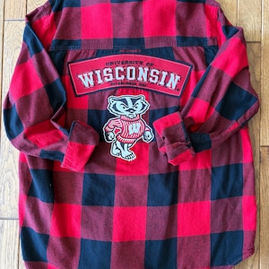 May include: A long-sleeve button-down shirt in a red and black buffalo plaid pattern. The back features a red patch with the text "UNIVERSITY OF WISCONSIN ESTABLISHED 1848" and a graphic of a badger mascot wearing a red sweater with a white W.
