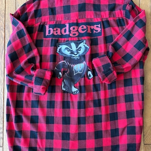 May include: A long-sleeve button-down shirt featuring a red and black buffalo plaid pattern. The back displays the word "badgers" in red text above a graphic illustration of a badger mascot wearing a dark sweater with a white letter W.