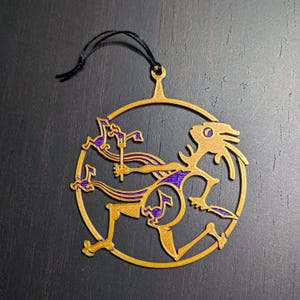 May include: Gold-colored metal ornament shaped like a circle with a stylized figure inside. The figure is purple and has a long, flowing tail. The ornament has a black string attached to the top for hanging.