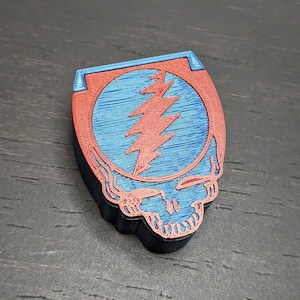 May include: A 3D-printed emblem featuring a red and blue Steal Your Face design. The emblem has a skull shape with a lightning bolt in the center. The colors are red, blue, and black.