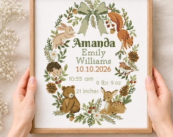 May include: A framed cross-stitch pattern featuring a woodland wreath with a rabbit, squirrel, hedgehog, bear, and deer. The text reads Amanda, Emily Williams, 10.10.2026, 10:55 am, 8 lbs 9 oz, and 21 inches. A sage green bow sits at the top of the wreath.