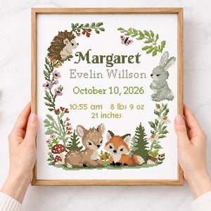 May include: A framed cross-stitch birth announcement with the name "Margaret" and birth details. The design includes woodland animals such as a hedgehog, rabbit, deer, and fox, surrounded by a floral wreath. The announcement is 53 cm in size.